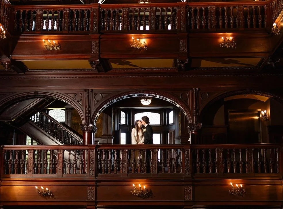 A wedding couple embraces on an ornate balcony staircase inside Grey Towers Castle, featuring richly carved dark wood,