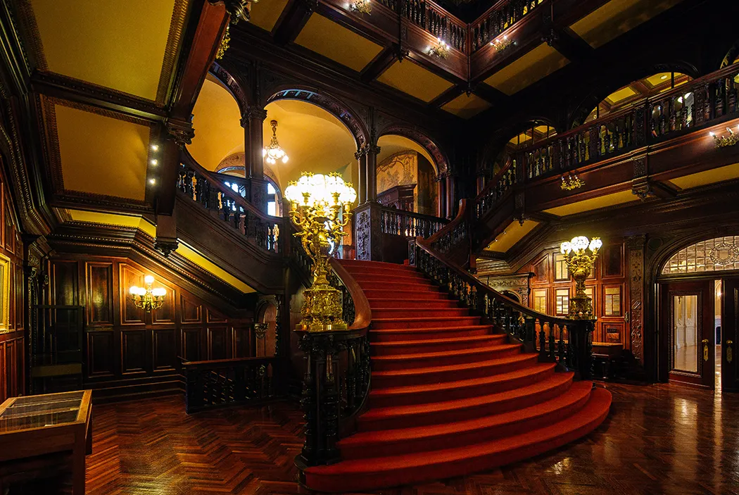 A grand staircase with red carpet, ornate décor, and rich dark woodwork, conveying elegance befitting a stately home.