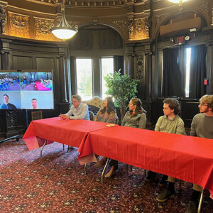 A panel of students from various STEM majors sitting at a table, with two on Zoom on a screen.