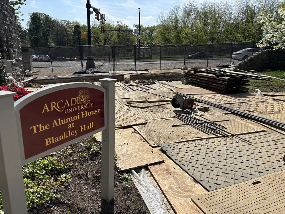 Construction materials, including planks, rods, plywood, and fencing, are arranged around the Rosemary D. Blankley ’57 Gatehouse Arch and Walkway near the Alumni House, indicating ongoing work in progress.