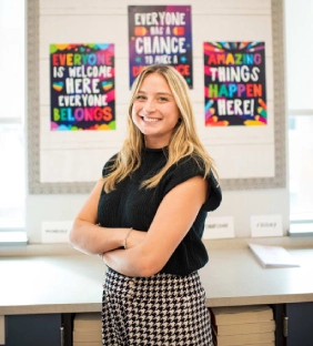 Honors Convocation 2025 award recipient Colleen Maria Hackendahl stands with her arms crossed and smiles in front of three posters. The first poster says: Everyone is welcome here, everyone belongs. The second poster says: Everyone has a chance to make a difference. The third poster says: Amazing things happen here!