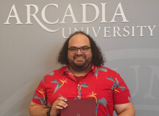 Honors Convocation 2025 award recipient Efrain Alex Nuñez stands in front of a wall with the Arcadia University text logo behind him and smiles for a photo while holding a dark red folder.