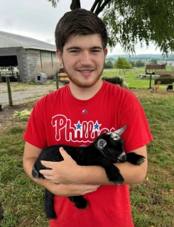 Honors Convocation 2025 award recipient Jason Robert Small stands at a goat pen holding a baby goat and smiles.