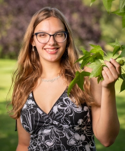 Honors Convocation 2026 award recipient Lucy Obrzut stands outside, holding her left hand up to hold a small tree branch with leaves on it, and smiles.