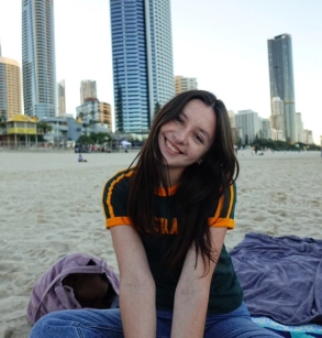 Honors Convocation 2025 award recipient Samantha Silverglade sits on a towel on a beach and smiles with large buildings visible in the background.