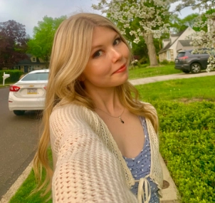 Honors Convocation 2025 award recipient Trisha McElhaugh stands on a sidewalk outside and tilts her head to the right while taking a selfie.