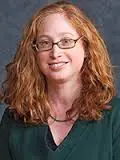 A headshot of a woman with red hair, glasses, and a green shirt