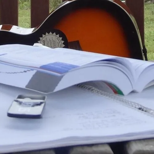 A guitar is propped on its side on a porch with an open book and notebook in front of it.
