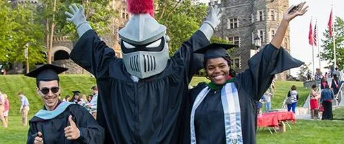 Two students in graduation caps and gowns stands with Arcadia knight mascot
