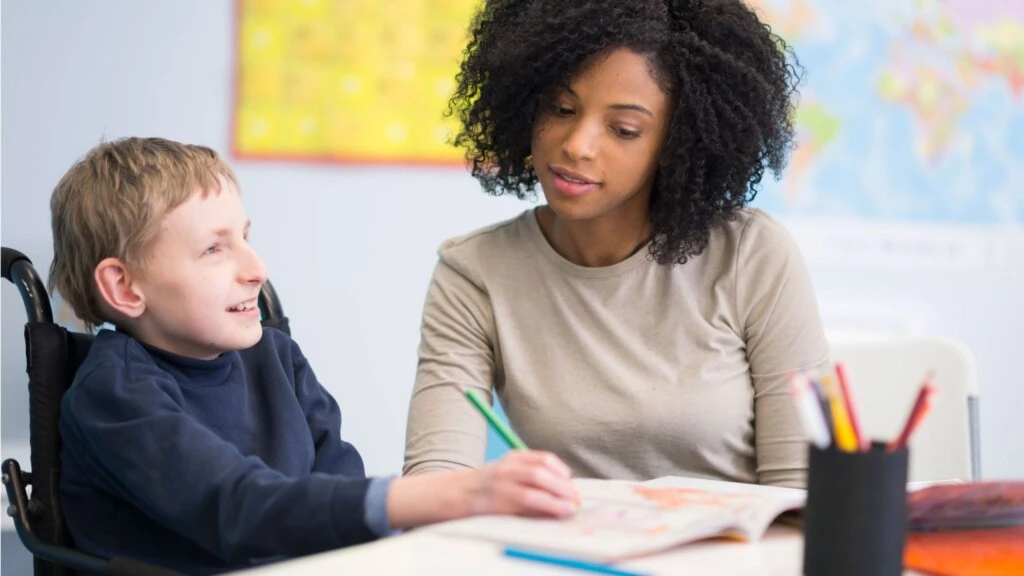 A teacher smiles while a young student draws in coloring book.