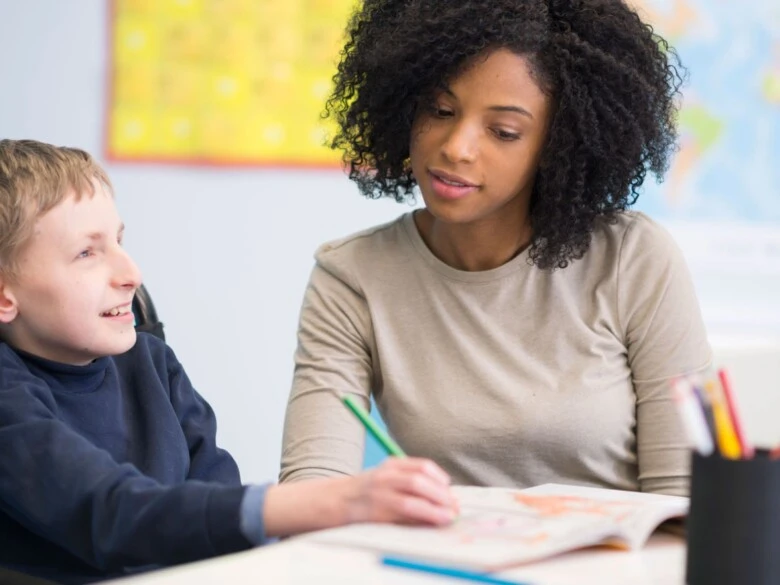 A teacher smiles while a young student draws in coloring book.