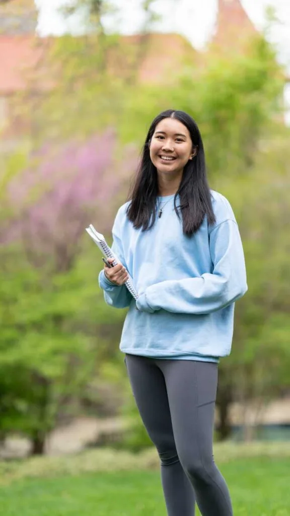 Smiling student stands on the lawn holding a book.