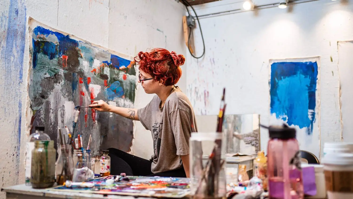 Student uses palette knife to spread blue, red, orange, black, and white oil paint on a canvas mounted on the studio wall.