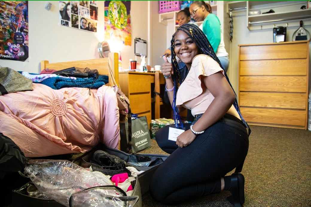 A student kneeling on the floor, smiling and holding her thumb up at the camera as she moves her clothing and shoes into her dorm on move-in day. Two students are in the background helping her move.
