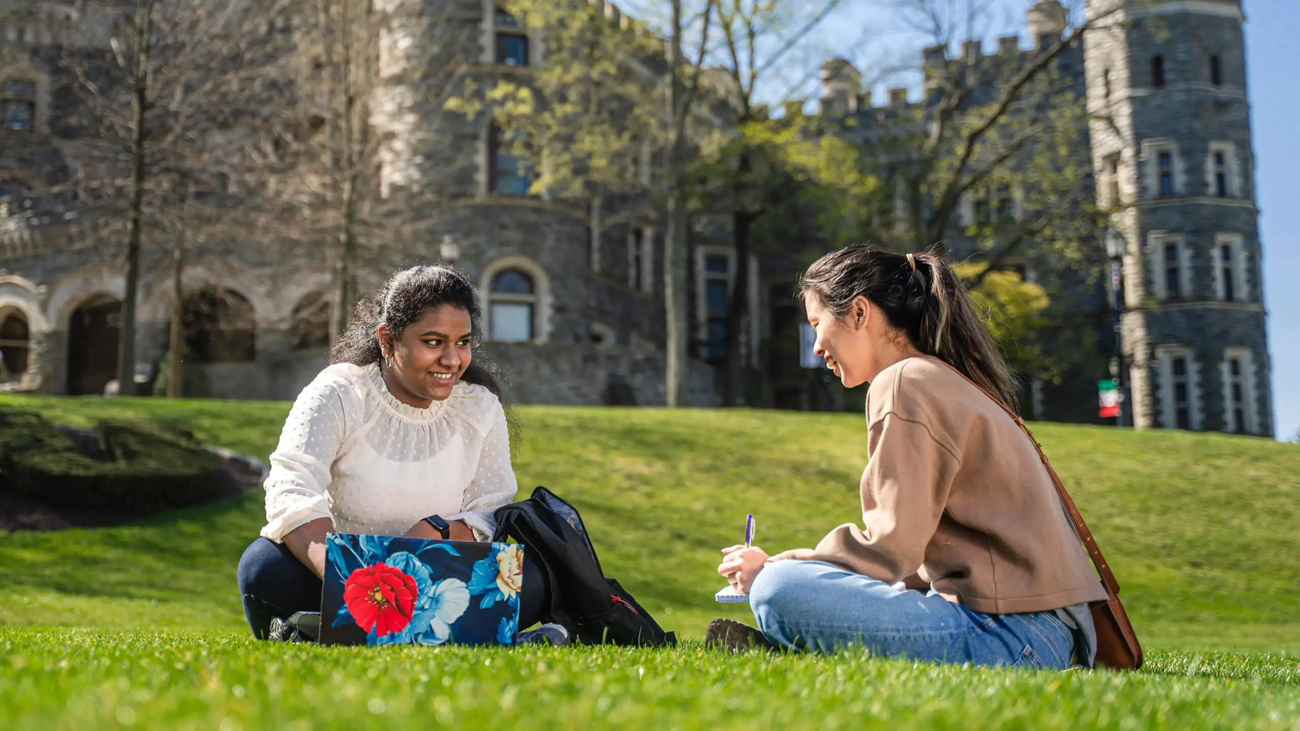 Two Arcadia University students smiling and sitting on the lawn in front of Grey Towers. The student on the left is using a laptop and the student on the right is writing notes in a notebook.