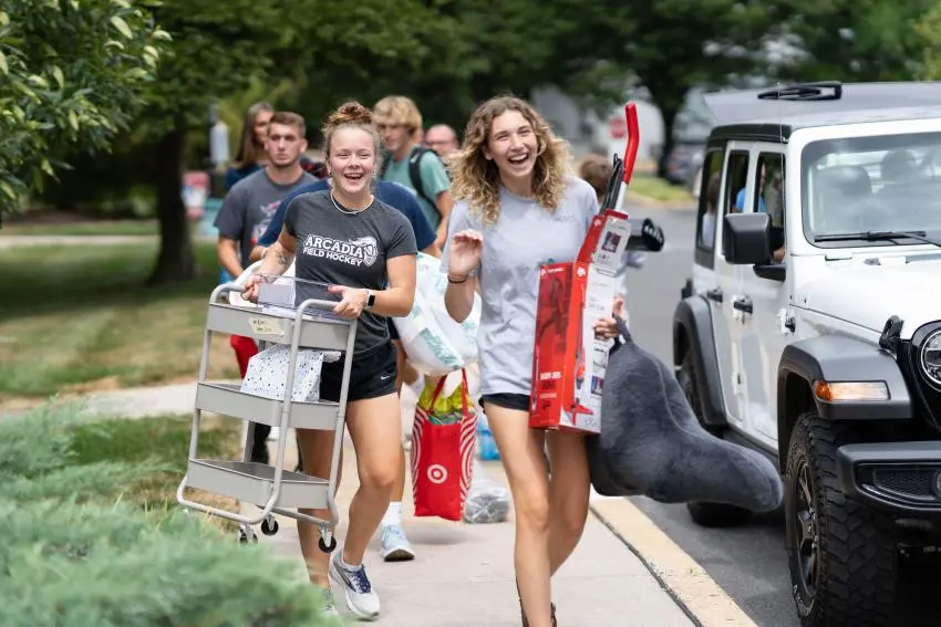A group of students walking down a street carrying groceries and organizers and a pillow