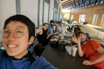 Selfie of a student at a commons dining area with others in the background waving
