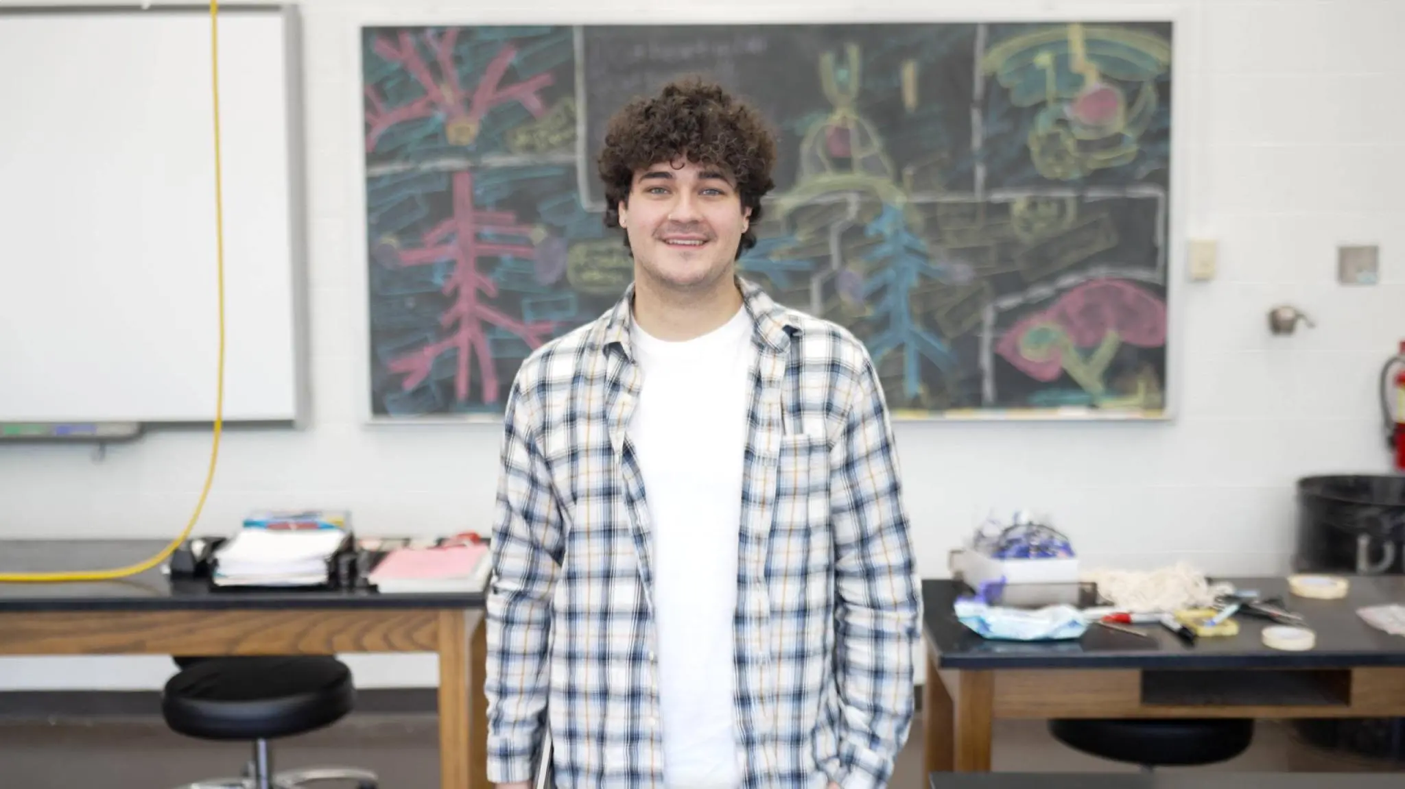 Student Jayson Cortez smiles at the camera while standing in front of a classroom blackboard