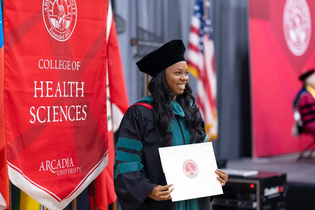 A graduate holds their diploma in front of the College of Health Sciences Banner 