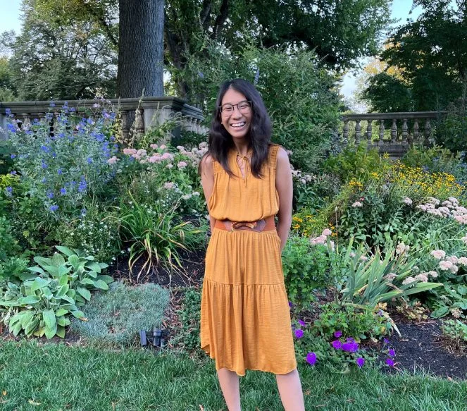 A female student with long dark hair stands in a garden smiling wearing a golden dress with a brown belt