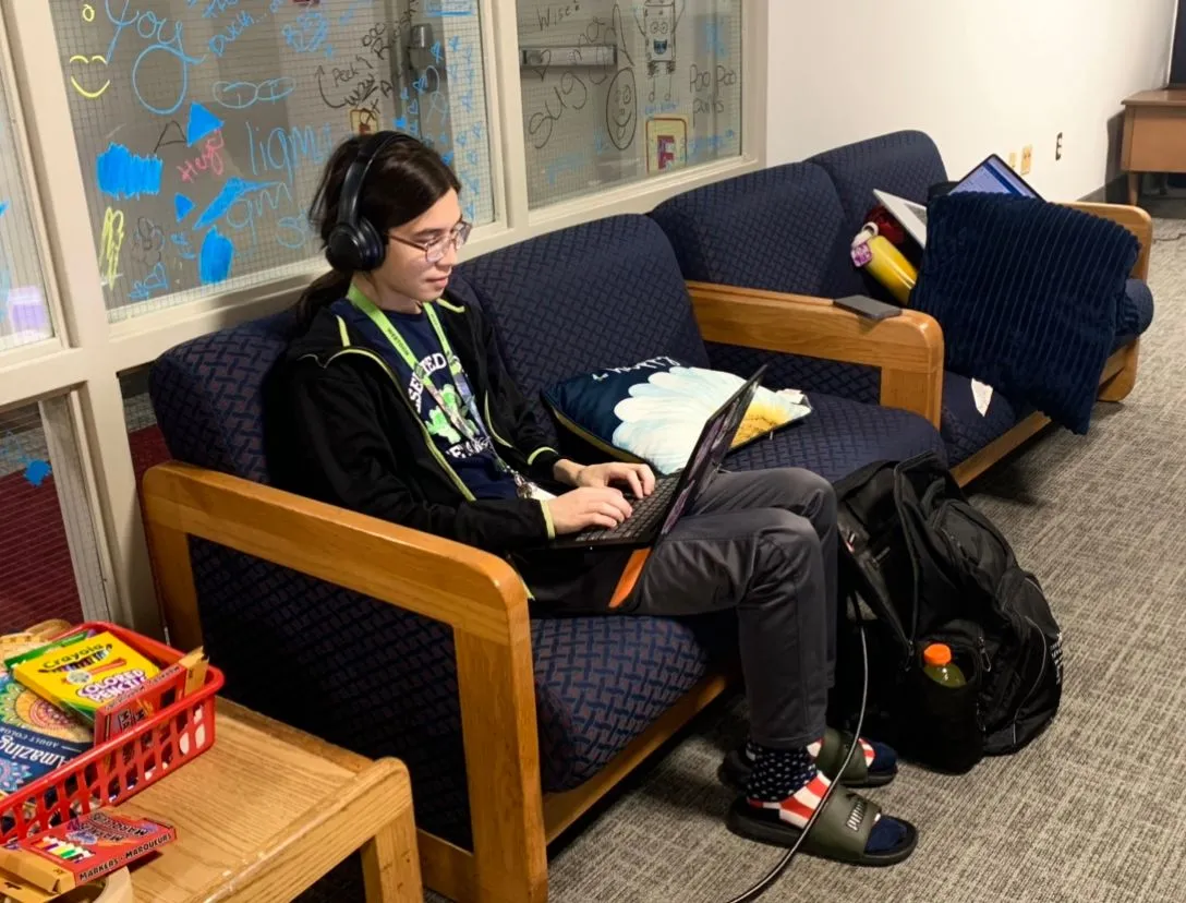 A student sits on a lounge couch wearing headphones and typing on a laptop