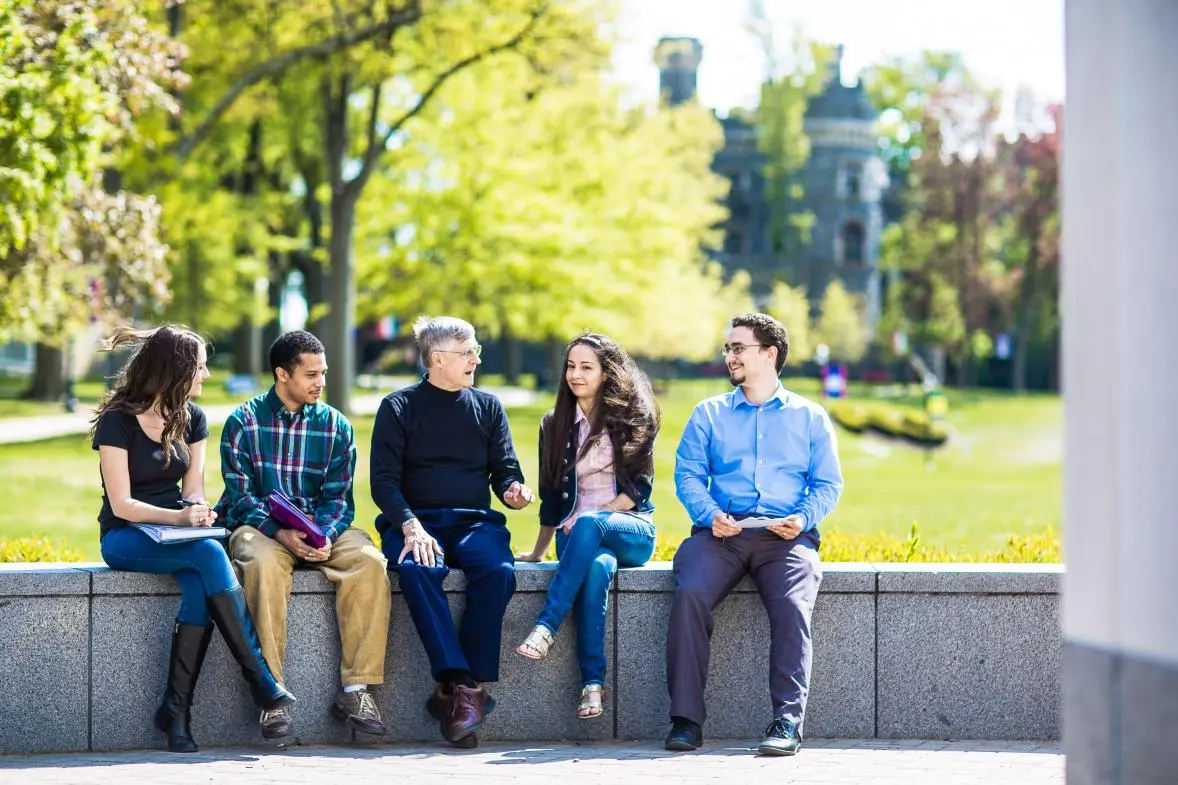 Students and faculty sit on a wall on campus and talk.