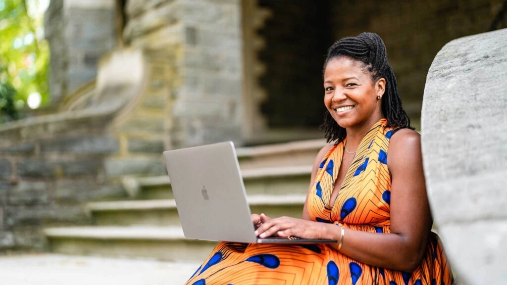 A Black woman in a colorful dress working on her Apple laptop and smiling into the camera.