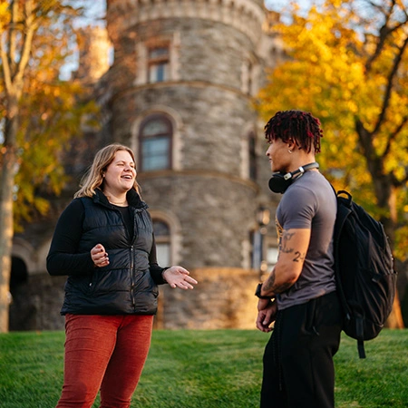 Two students have a chat in front of the castle on campus on a leafy fall day.