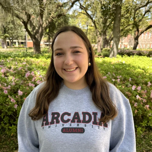 Young woman with brown hair in a grey Arcadia sweatshirt. Standing in front of a tree.