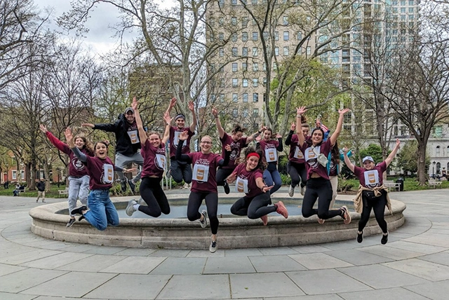 A group of PA students jump and wave their hands in the air in front of historical buildings in a park.
