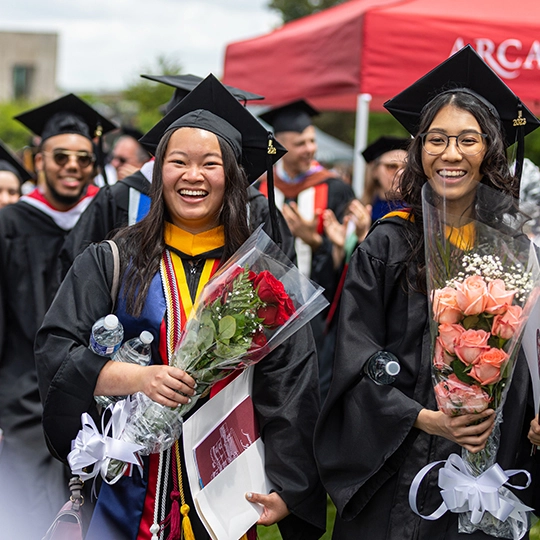 Two undergraduate students smile during commencement excercises and hold bunches of flowers.